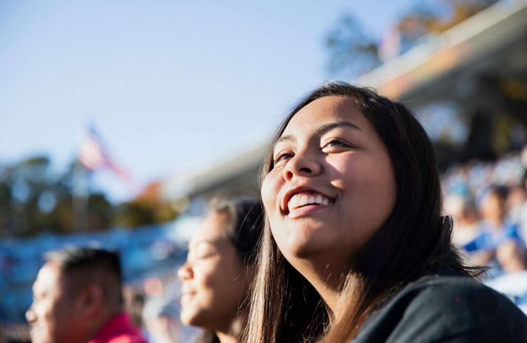 A smiling student sits in the stands at a football game looking out into the horizon