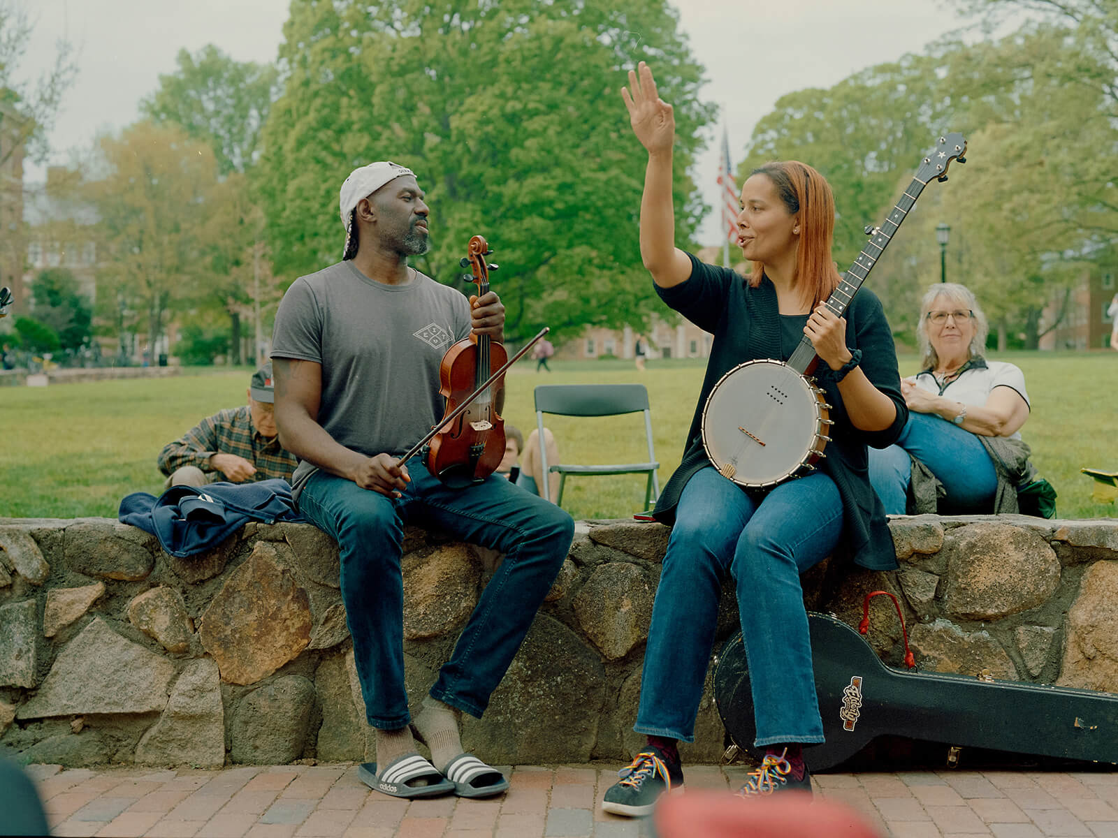 Rhiannon Giddens sitting on a stone wall on Polk Place while holding a banjo, next to a musician holding a fiddle