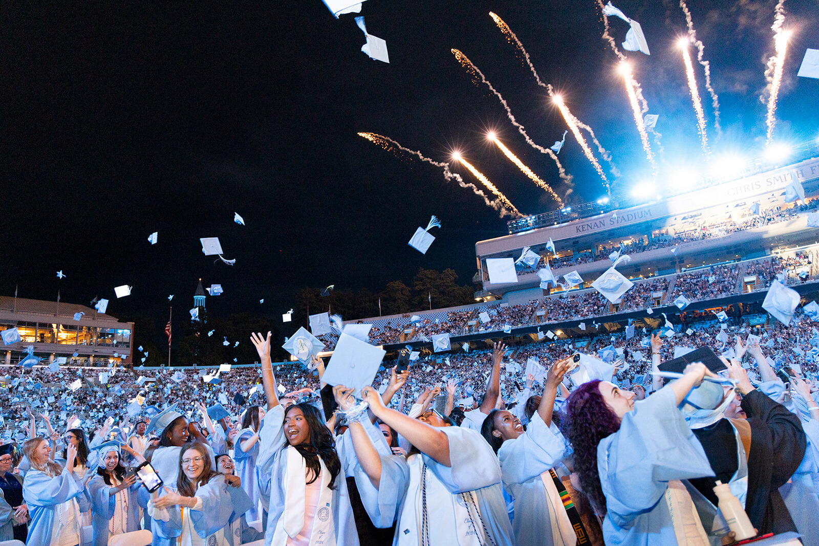 Carolina graduates, in graduation regalia, throwing their caps into the air in Kenan Stadium while fireworks go off in the background