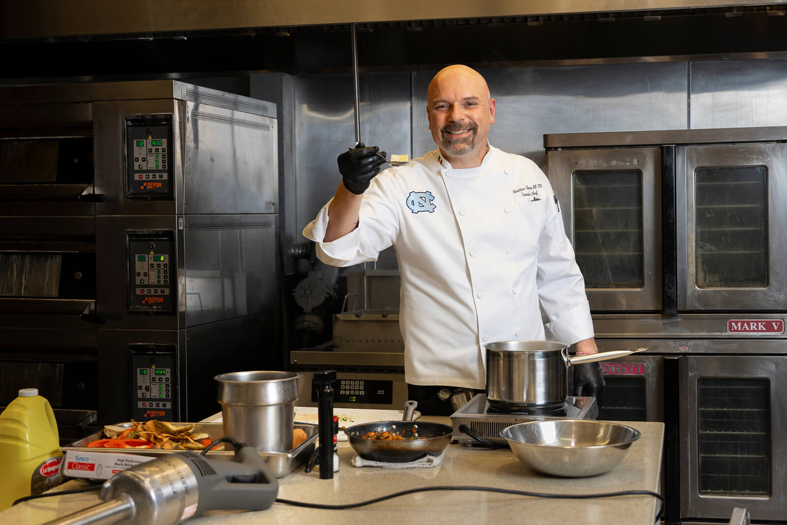 Christian Chiron standing in a kitchen in a chef’s coat while smiling and holding up ingredients on a spoon