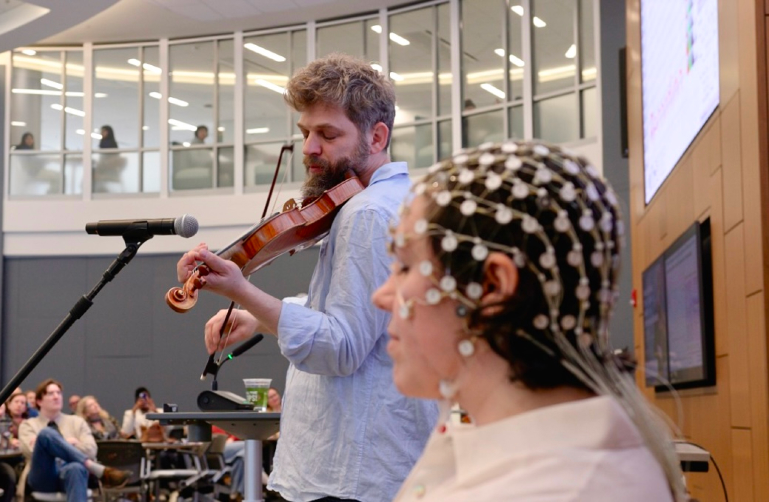 Johnny Gandelsman plays violin in front of a microphone in the background; a woman wears tACS headgear listens to the music in the foreground