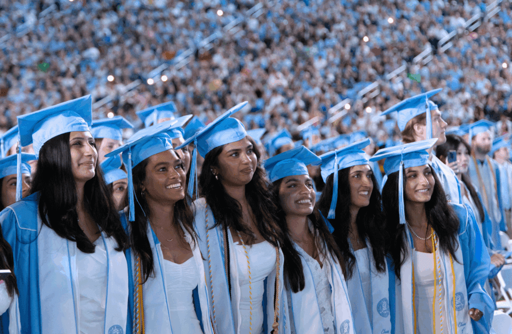 Six graduates smiling in a line, standing up.