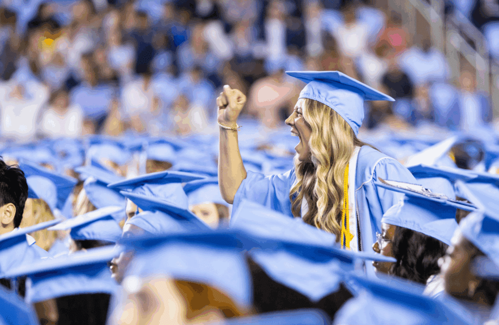 A singular student standing up, clenching their fist and yelling with excitement in a crowd of graduates.