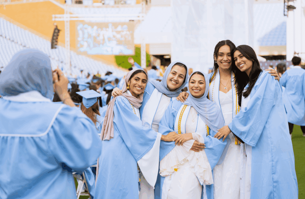 A group of five Carolina graduates smile for a phone camera.