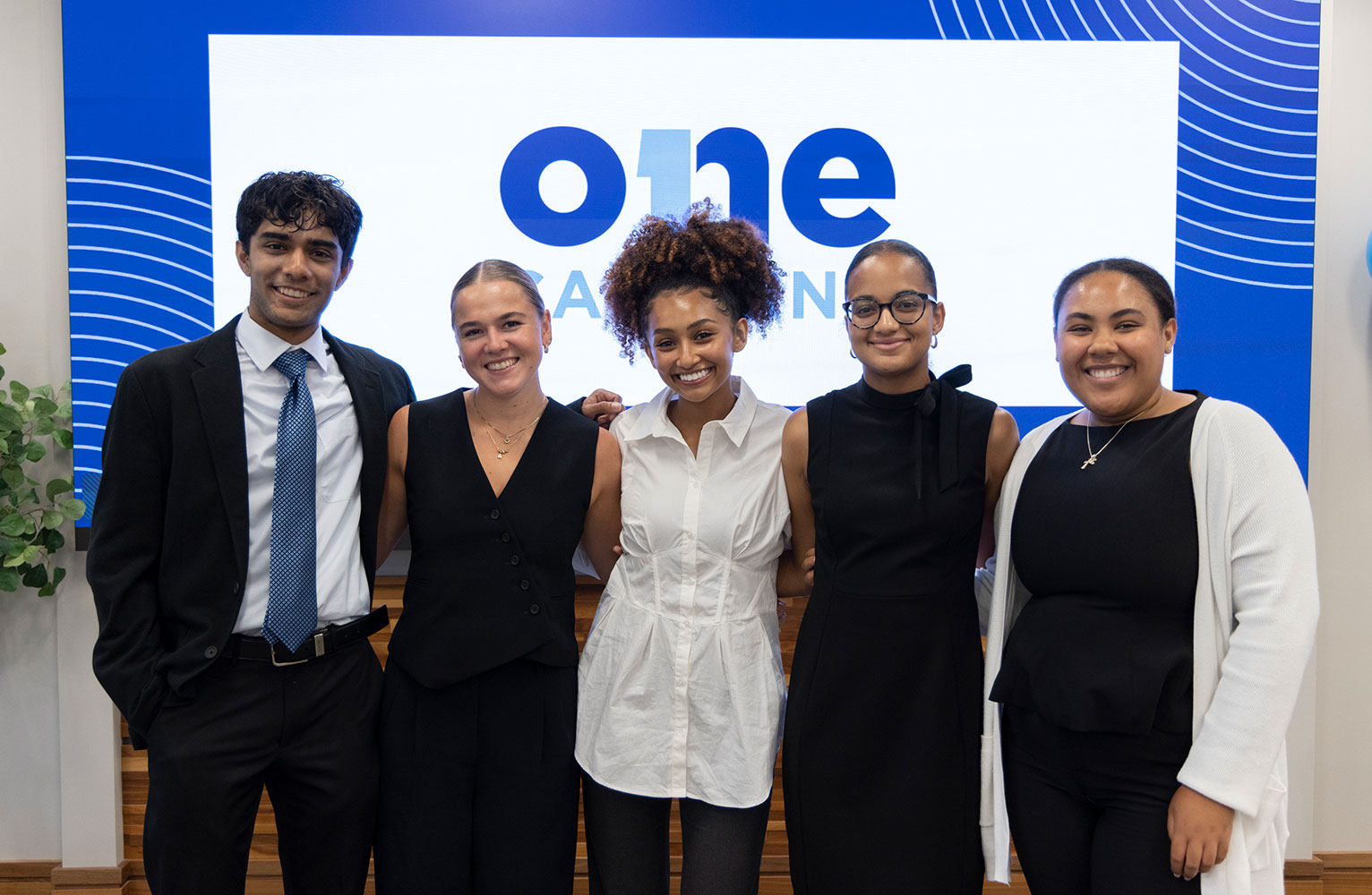 Five Carolina students stand side by side and face the camera with the OneCarolina logo on a screen in the background
