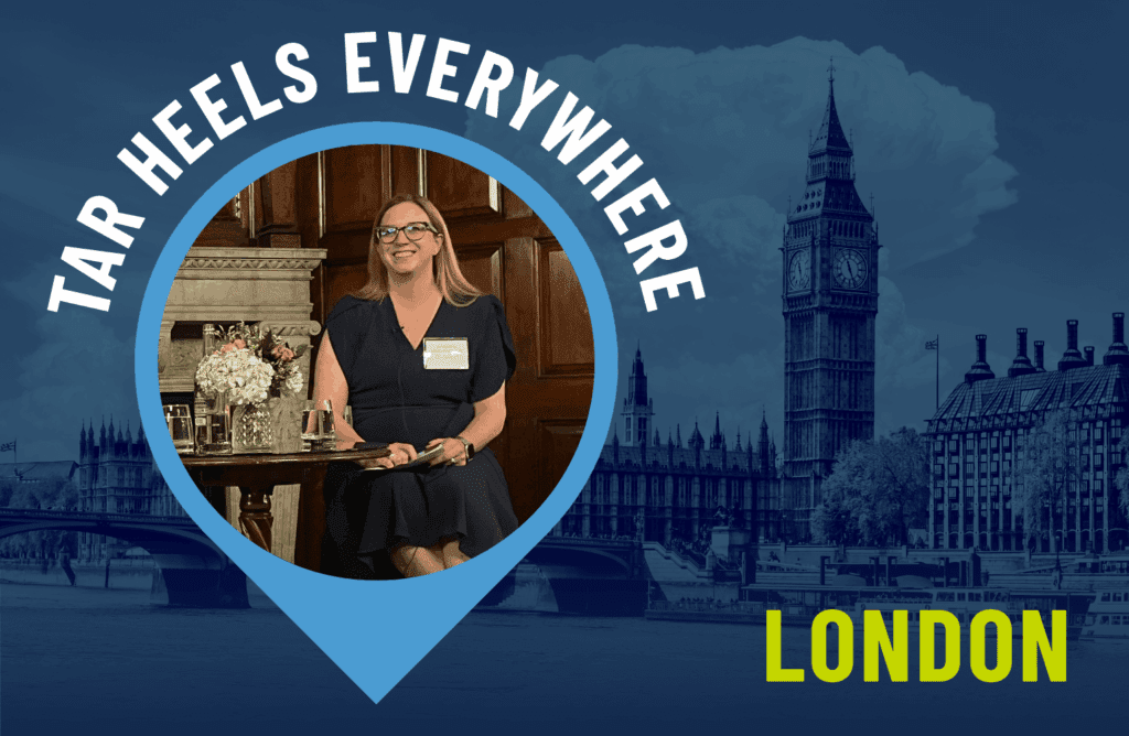 Photo of a smiling woman seated indoors, wearing glasses and a dark dress, with flowers on the table beside her. She is framed within a blue location pin graphic labeled ‘Tar Heels Everywhere.’ In the background, a stylized image of London’s Big Ben and the Houses of Parliament appears, with the word ‘London’ in bold yellow text.