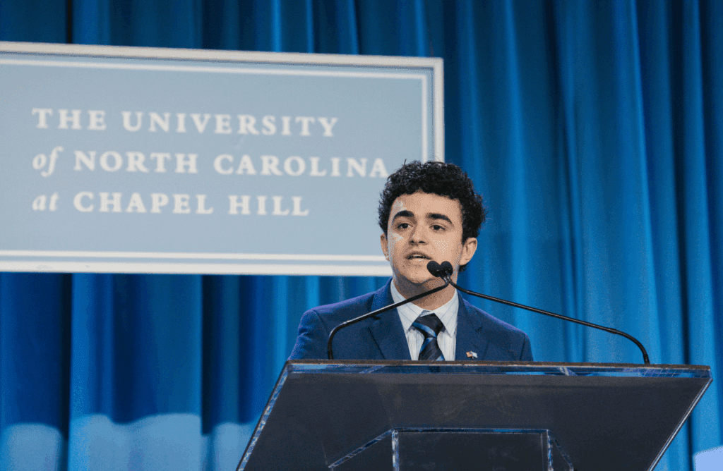 Man in blue suit speaking into a microphone on stage with blue curtains behind him.