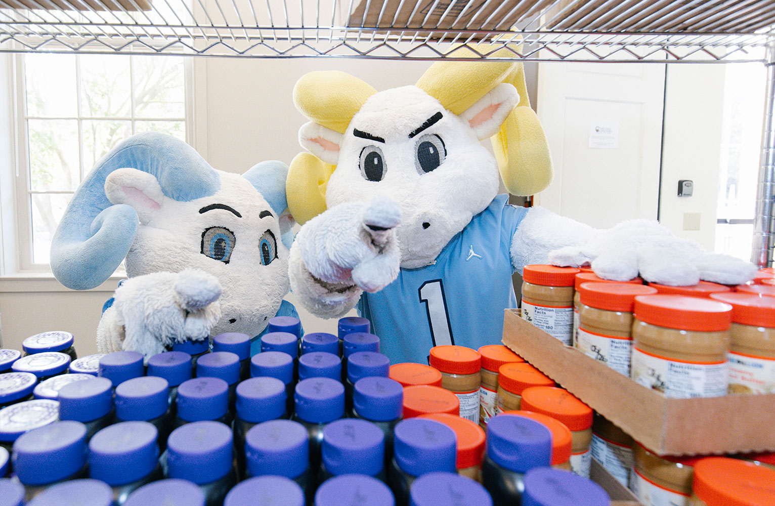 RJ and Rameses point at the camera in a campus food pantry, with jars of peanut butter in the foreground.