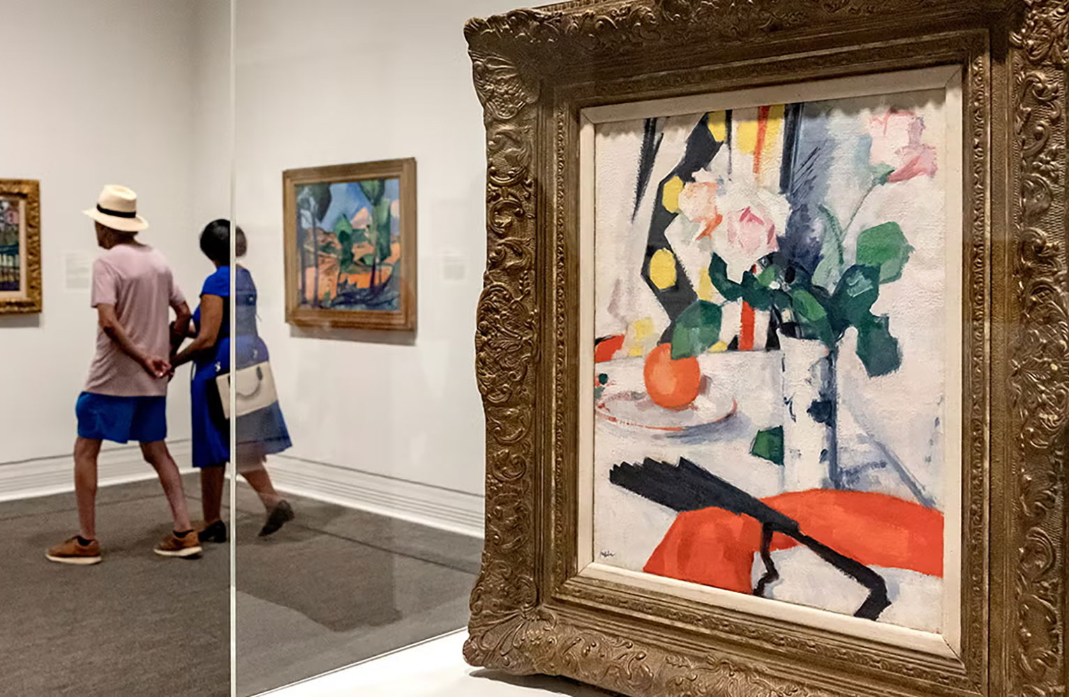 The Painting "Still Life of Tulips in a Chinese Vase with a Black Fan" in the foreground with a couple holding hands viewing other artworks in the "Color Triumphant" exhibit in the background