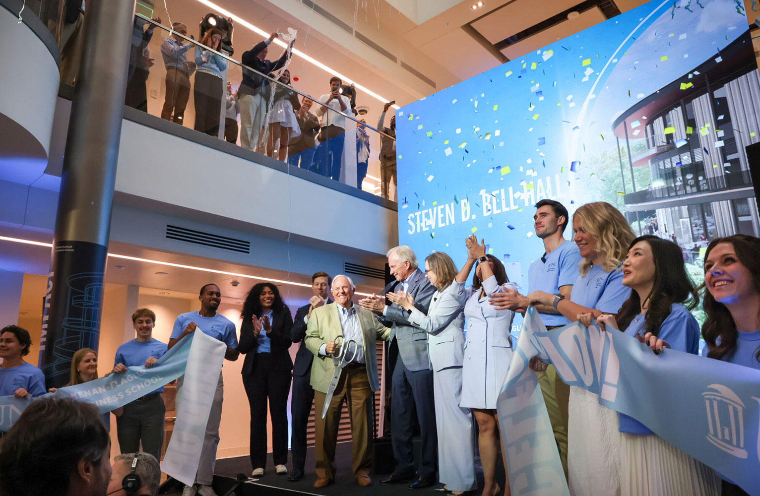 A group of stakeholders clap joyfully in front of a Bell Hall sign at the opening of Bell Hall.