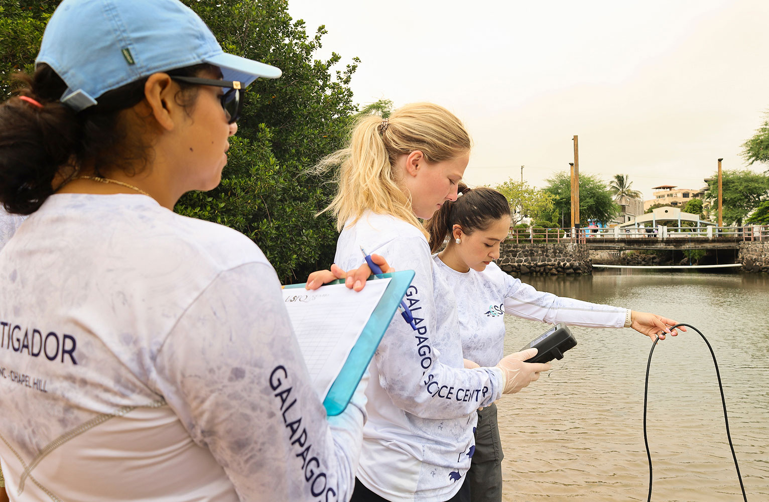 Three women stand on a shoreline with water testing equipment.