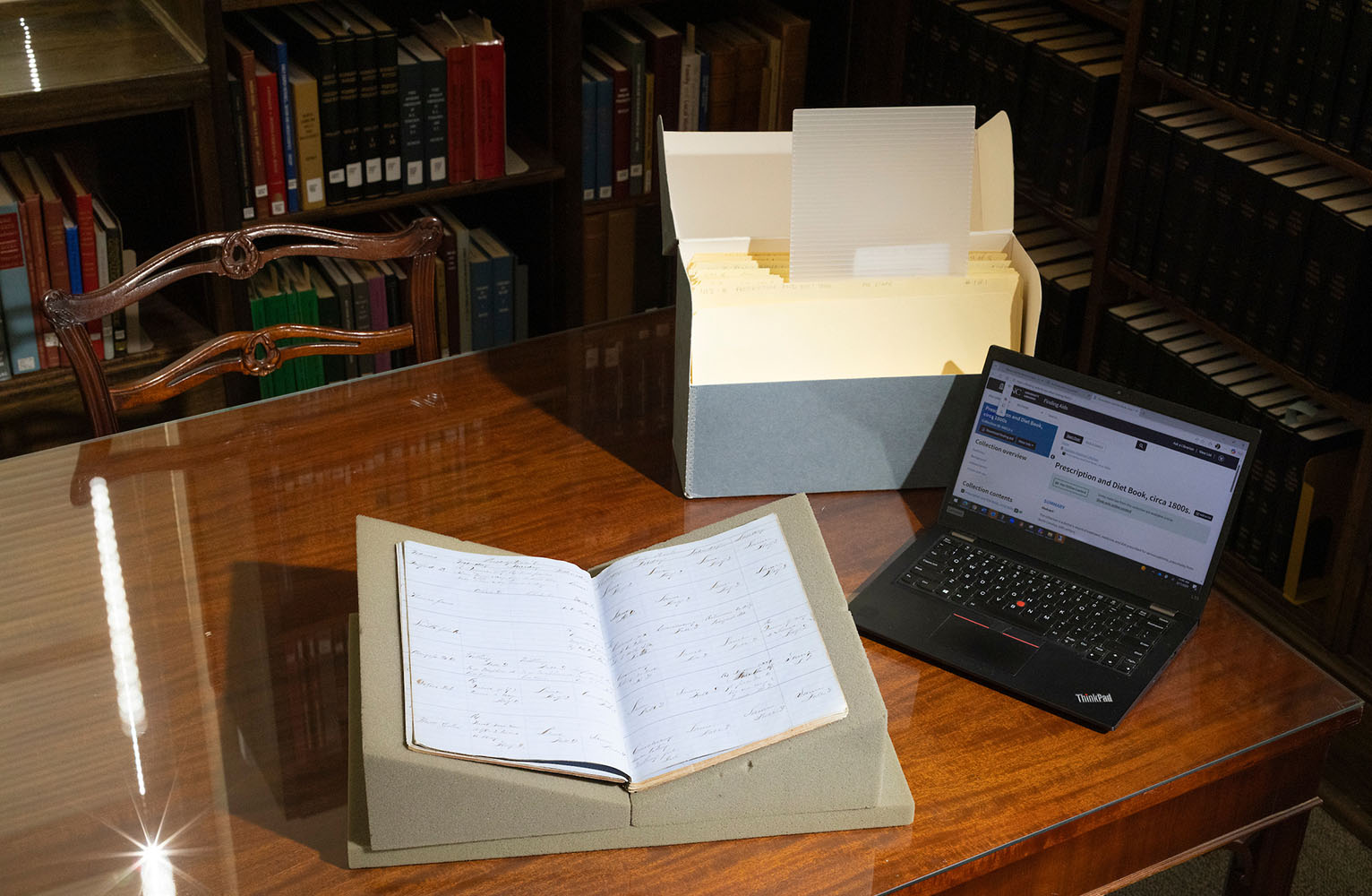 A splayed-open daybook, open laptop and box of files are arranged on a wooden table in a reading room at Wilson Special Collections Library