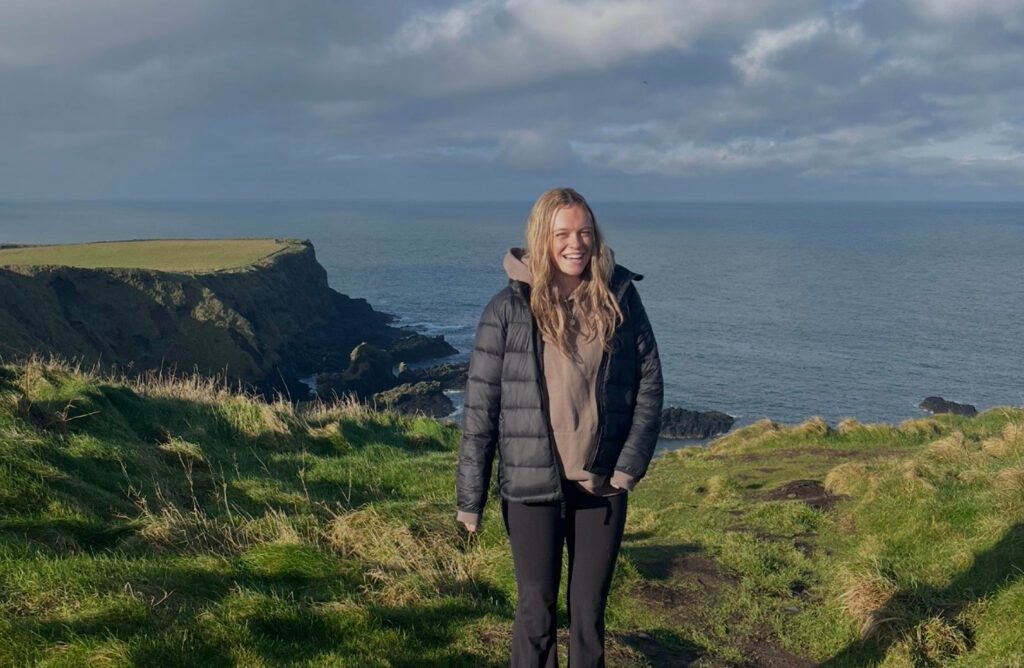 Caroline Robinson stands on a grassy plateau overlooking water in Ireland.