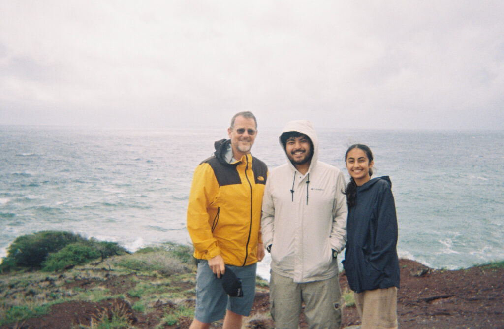 Patrick Davison, Aayas Joshi and Keya Mahajan stand on a cliff overlooking the ocean on a cloudy day in Hawaii.