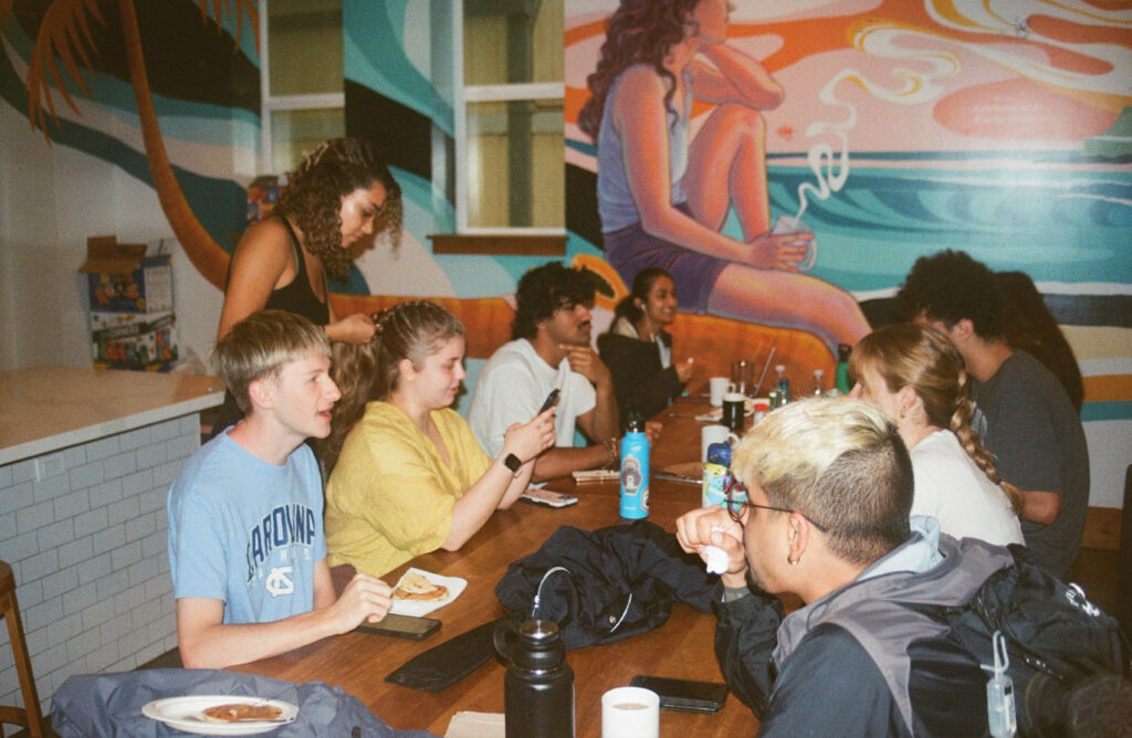 Students convene at a dining table in the hostel, with students pictured eating, talking and braiding one another's hair.