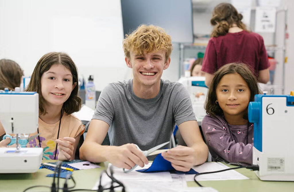 Austin Roebuck smiles facing the camera with young girls on either side of him sewing.