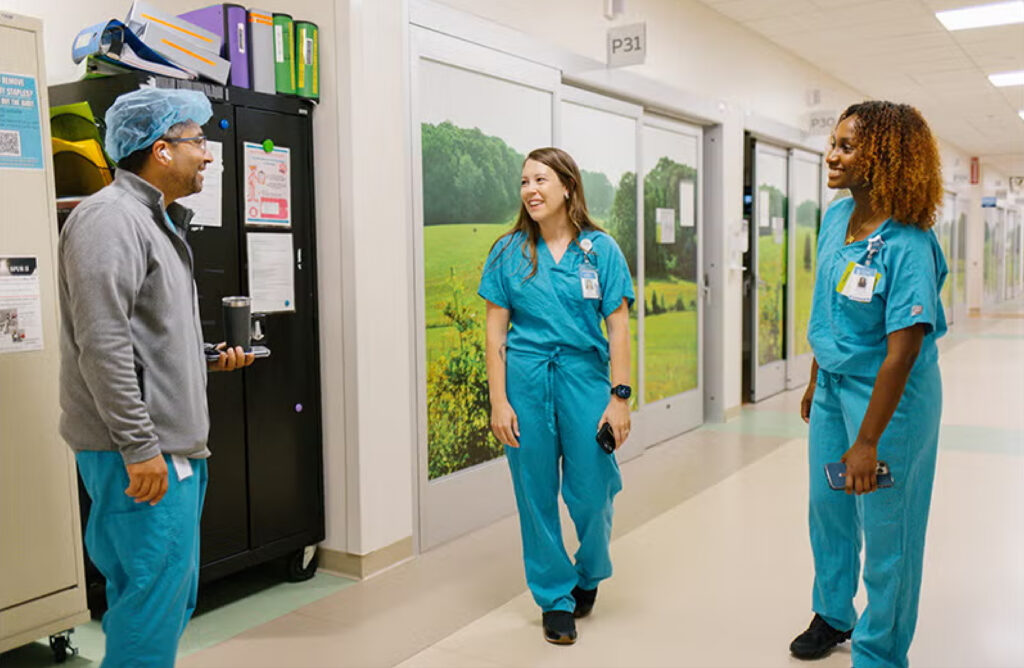 Three healthcare workers in teal scrubs converse in a hospital hallway adorned with nature-themed wall murals.